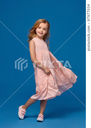 Fashionable little girl in a pink dress is posing on a blue background. 97977034