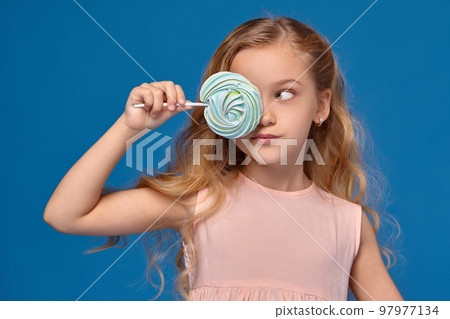 Fashionable little girl in a pink dress, with a candy in her hands, standing on a blue background. Fashionable little girl in a pink dress, with a candy in her hands, standing on a blue background. 97977134
