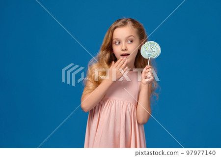 Fashionable little girl in a pink dress, with a candy in her hands, standing on a blue background. Fashionable little girl in a pink dress, with a candy in her hands, standing on a blue background. 97977140