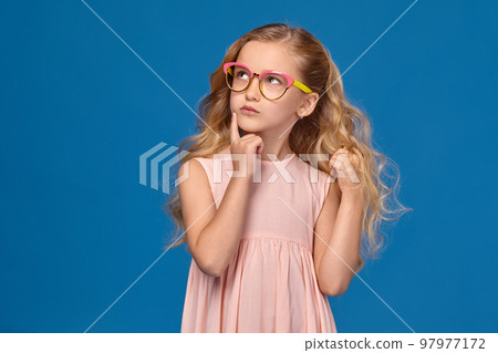 Fashionable little girl in a pink dress and glasses is standing on a blue background. Fashionable little girl in a pink dress and glasses is standing on a blue background. 97977172