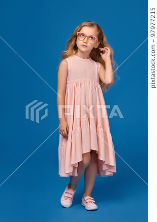 Fashionable little girl in a pink dress and glasses is standing on a blue background. 97977215