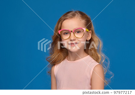 Fashionable little girl in a pink dress and glasses, with a candy in her hands, standing on a blue background. 97977217