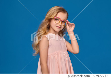 Fashionable little girl in a pink dress and glasses is standing on a blue background. 97977218