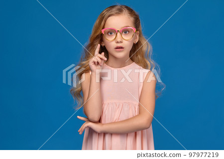 Fashionable little girl in a pink dress and glasses is standing on a blue background. 97977219