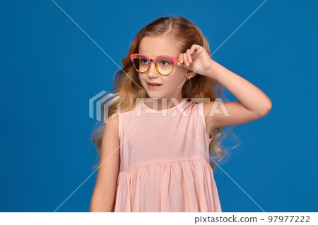 Fashionable little girl in a pink dress and glasses is standing on a blue background. Fashionable little girl in a pink dress and glasses is standing on a blue background. 97977222