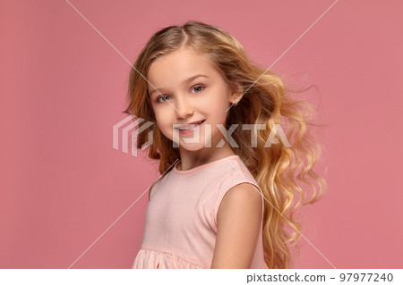 Little girl with a blond curly hair, in a pink dress is posing for the camera 97977240