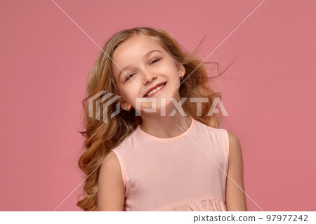 Little girl with a blond curly hair, in a pink dress is posing for the camera 97977242