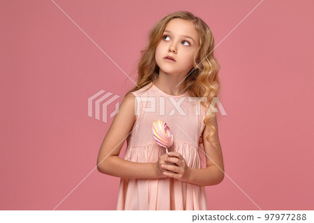 Little girl with a blond curly hair, in a pink dress is posing with a candy 97977288