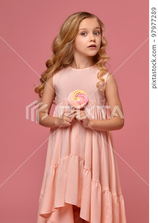 Little girl with a blond curly hair, in a pink dress is posing with a candy Little girl with a blond curly hair, in a pink dress is posing with a candy 97977289