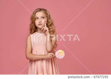 Little girl with a blond curly hair, in a pink dress is posing with a candy Little girl with a blond curly hair, in a pink dress is posing with a candy 97977295