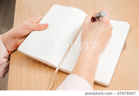 Close up of a woman's hands writing in a notebook placed on a wooden table, home office and work concept, plans and business meeting Close up of a woman's hands writing in a notebook placed on a wooden table, home office and work concept, plans and business meeting 97978097