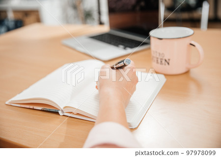 Close up of a woman's hands writing in a notepad placed on a wooden table, laptop, home office and work concept, plans and business meeting Close up of a woman's hands writing in a notepad placed on a wooden table, laptop, home office and work concept, plans and business meeting 97978099