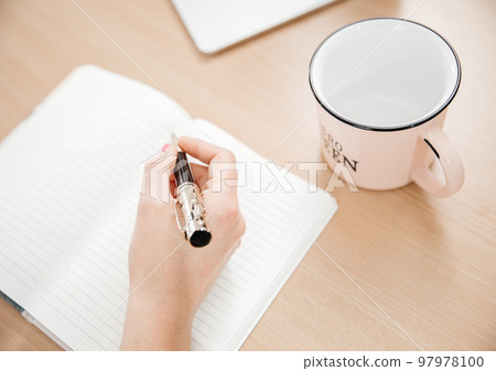 Close up of a woman's hands writing in a notepad placed on a wooden table, cup for tea, home office concept Close up of a woman's hands writing in a notepad placed on a wooden table, cup for tea, home office concept 97978100