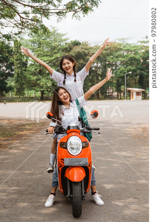 Two high school girls riding motorbikes standing while celebrating graduation Two high school girls riding motorbikes standing while celebrating graduation 97980802