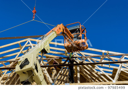 During construction of rafters an unfinished wooden house crane holds wood trusses beams to roof During construction of rafters an unfinished wooden house crane holds wood trusses beams to roof 97985499