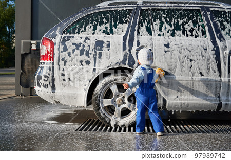 Rear view of toddler boy in jumpsuit and cap, standing with sponge in his hands near side door of car body covered with soap suds. Child rubbing soap suds, taking part in cleaning auto at the car wash Rear view of toddler boy in jumpsuit and cap, standing with sponge in his hands near side door of car body covered with soap suds. Child rubbing soap suds, taking part in cleaning auto at the car wash 97989742