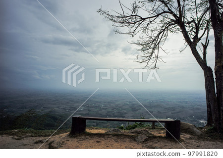 Empty wooden bench under the tree on the mountain with landscape and cloudy sky in background Empty wooden bench under the tree on the mountain with landscape and cloudy sky in background 97991820