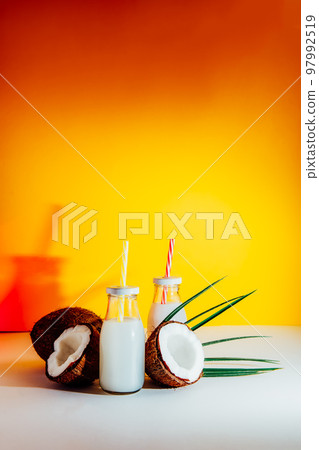 Bottles with coconut non-dairy milk drink and fresh coconuts on bright yellow background with color light. Trendy coctails. Healthy vegan plant base coconut milk. Vertical. Selective focus. Copy space Bottles with coconut non-dairy milk drink and fresh coconuts on bright yellow background with color light. Trendy coctails. Healthy vegan plant base coconut milk. Vertical. Selective focus. Copy space 97992519