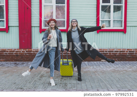 Two cheerful funny tourist women smiling and fool around and with suitcases on city street in autumn or spring time - travel and vacation concept 97992531
