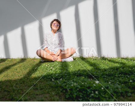 Smiling woman in colorful sunglasses has a rest on lawn in urban park. Nature in town. Relax outdoors after work. Summer vibes. Smiling woman in colorful sunglasses has a rest on lawn in urban park. Nature in town. Relax outdoors after work. Summer vibes. 97994114
