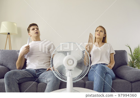 Young couple who are tired from extremely hot summer weather use electric fan at home 97994360