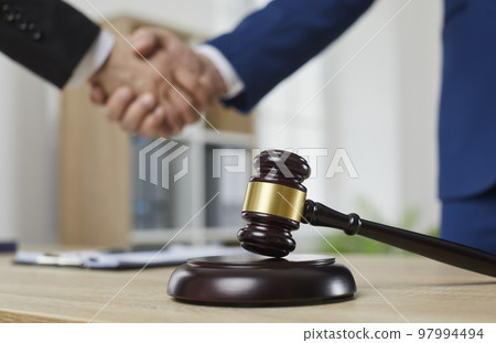 Close-up of a wooden court hammer on stand on table in the courtroom against the background of handshake. 97994494