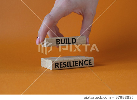 Build resilience symbol. Wooden blocks with words Build resilience. Businessman hand. Beautiful orange background. Business and Build resilience concept. Copy space. 97998855