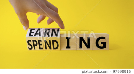 Earning and Spending symbol. Businessman hand. Turned cubes with words Earning and Spending. Beautiful yellow background. Business and Earning and Spending concept. Copy space Earning and Spending symbol. Businessman hand. Turned cubes with words Earning and Spending. Beautiful yellow background. Business and Earning and Spending concept. Copy space 97998916