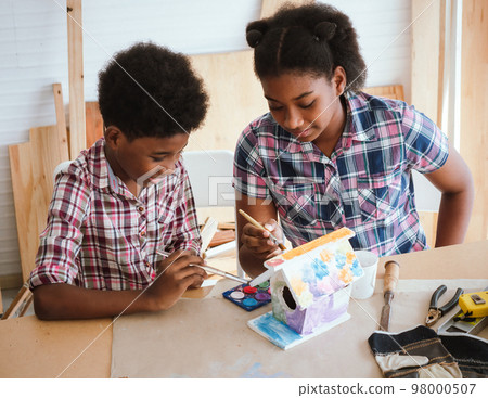 Two African American brothers and sisters help each other make wooden signs to celebrate the Christmas season in the workshop.. Two African American brothers and sisters help each other make wooden signs to celebrate the Christmas season in the workshop.. 98000507