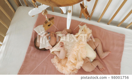 Top view of baby girl in dress and bow headband looking at toys on hanging mobile 98001756
