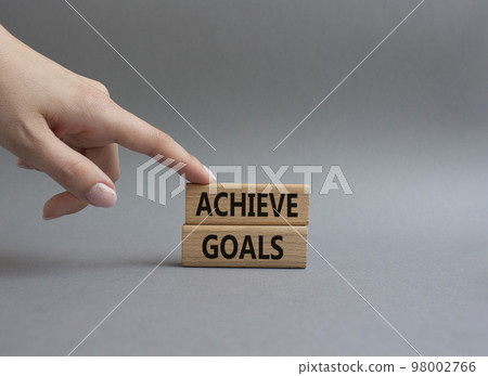 Achieve goals symbol. Concept words achieve goals on wooden blocks. Beautiful grey background. Businessman hand. Business and achieve goals concept. Copy space. Achieve goals symbol. Concept words achieve goals on wooden blocks. Beautiful grey background. Businessman hand. Business and achieve goals concept. Copy space. 98002766