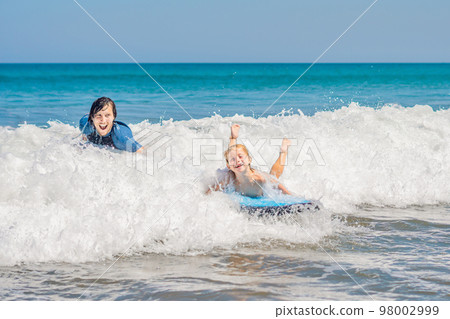 Father teaching his young son how to surf in the sea on vacation or holiday. Travel and sports with children concept 98002999