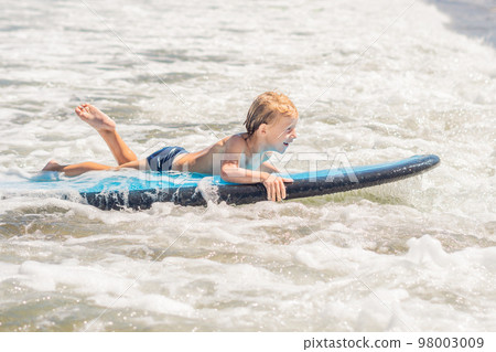 Happy baby boy - young surfer ride on surfboard with fun on sea waves. Active family lifestyle, kids outdoor water sport lessons and swimming activity in surf camp. Summer vacation with child Happy baby boy - young surfer ride on surfboard with fun on sea waves. Active family lifestyle, kids outdoor water sport lessons and swimming activity in surf camp. Summer vacation with child 98003009
