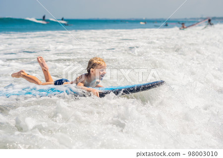 Happy baby boy - young surfer ride on surfboard with fun on sea waves. Active family lifestyle, kids outdoor water sport lessons and swimming activity in surf camp. Summer vacation with child 98003010