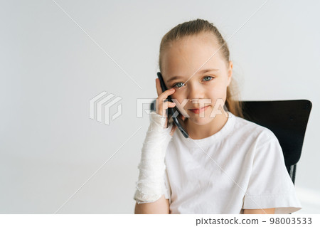 Medium shot portrait of cute little girl with broken arm wrapped in plaster bandage talking smartphone, looking at camera, sitting chair on white background. Concept of child insurance and healthcare. 98003533