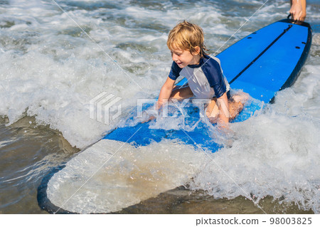 Happy baby boy - young surfer ride on surfboard with fun on sea waves. Active family lifestyle, kids outdoor water sport lessons and swimming activity in surf camp. Summer vacation with child Happy baby boy - young surfer ride on surfboard with fun on sea waves. Active family lifestyle, kids outdoor water sport lessons and swimming activity in surf camp. Summer vacation with child 98003825