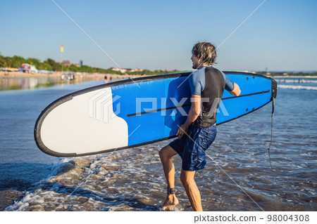 Handsome sporty young surfer posing with his surfboard under his arm in his wetsuit on a sandy tropical beach 98004308
