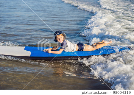 Happy baby boy - young surfer ride on surfboard with fun on sea waves. Active family lifestyle, kids outdoor water sport lessons and swimming activity in surf camp. Summer vacation with child Happy baby boy - young surfer ride on surfboard with fun on sea waves. Active family lifestyle, kids outdoor water sport lessons and swimming activity in surf camp. Summer vacation with child 98004319