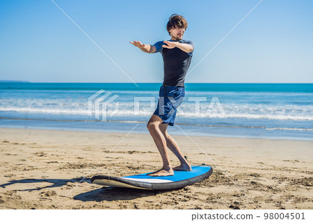 Young man surfer training before go to lineup on a sand beach. Learning to surf. Vacation concept. Summer holidays. Tourism, sport 98004501