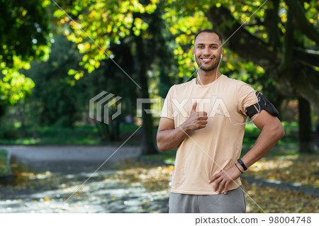 Portrait of successful man in park hispanic man with headphones listening to music and audiobooks online podcasts, smiling and looking at camera while jogging and doing fitness exercise. 98004748