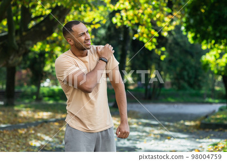 A man injured his shoulder during a fitness class, an African-American man injured himself while jogging in the park, stretches his arm and massages his sore muscles. A man injured his shoulder during a fitness class, an African-American man injured himself while jogging in the park, stretches his arm and massages his sore muscles. 98004759