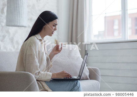 Upset young woman sits at home on the couch by the window with a laptop, reads bad news, received an email. Crying, wiping tears with a napkin. 98005446