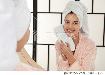 Young woman wiping face with towel near mirror in bathroom 98006338