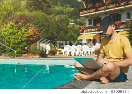 Young man working with laptop next to the pool during a sunny day. Attractive man working. Freelancer. Traveler.  98006384