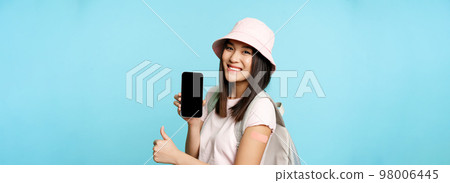 Smiling asian tourist shows arm with patch from covid-19 vaccination, showing coronavirus vaccine international certificate on smartphone app, thumbs up Smiling asian tourist shows arm with patch from covid-19 vaccination, showing coronavirus vaccine international certificate on smartphone app, thumbs up 98006445