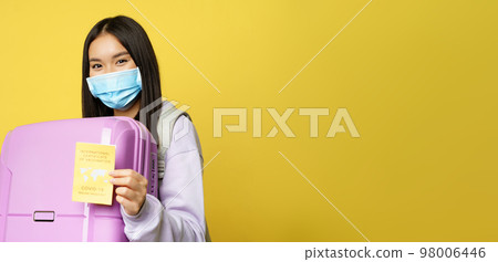 Covid-19 and tourism concept. Happy asian female tourist holding suitcase, showing international coronavirus vaccination certificate for travelling, yellow background Covid-19 and tourism concept. Happy asian female tourist holding suitcase, showing international coronavirus vaccination certificate for travelling, yellow background 98006446