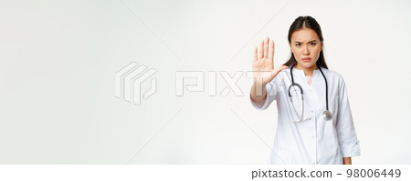 Asian doctor shows palm, stop gesture. Serious displeased female physician extend her arm, prohibit action, taboo sign, standing in uniform against white background Asian doctor shows palm, stop gesture. Serious displeased female physician extend her arm, prohibit action, taboo sign, standing in uniform against white background 98006449