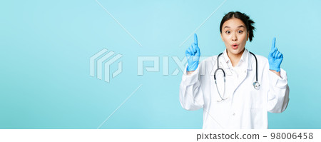 Excited asian medical worker, female doctor pointing fingers up, wearing sterile gloves and uniform, standing over blue background 98006458