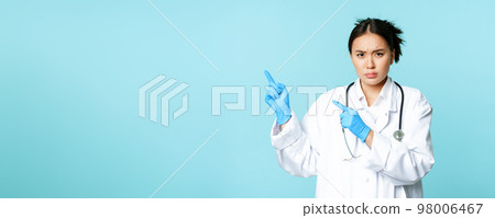 Angry female doctor or nurse, pointing fingers at upper left corner with disapproval, sulking and furrow eyebrows, standing in medical uniform, blue background 98006467