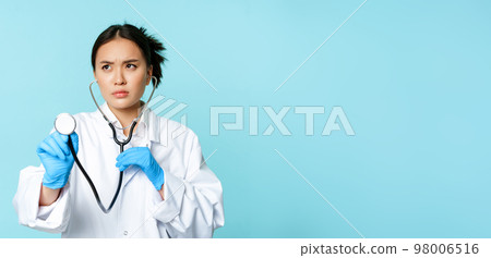 Image of serious medical worker, asian doctor listening to patient lungs with stethoscope, examining person, standing over blue background 98006516